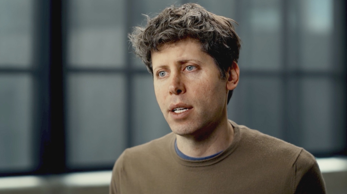 A man in a brown sweater talking to camera in an office