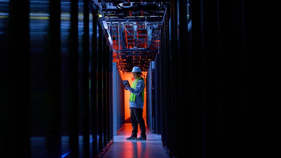 An engineer in a hard hat at the end of a dark server aisle lit red